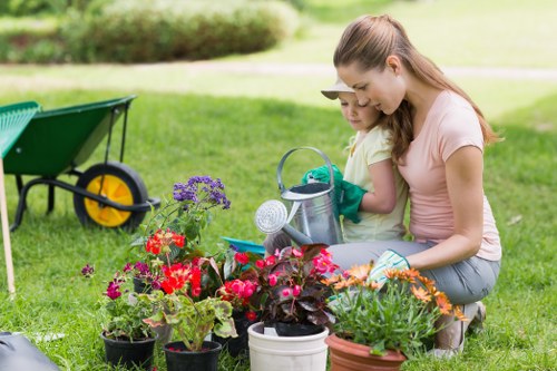 A woman and young girl are kneeling on a well-maintained grassy garden lawn in an outdoor space, engaging in gardening activities. The foreground features a collection of vibrant flowering plants in pots, including purple, red, and orange blooms, with some plants having lush green foliage. Behind them, a neat flower bed border separates the garden area from a background of trimmed hedges and trees, providing natural privacy. To the left, a green wheelbarrow with yellow handles rests on the grass, indicating ongoing garden work or planting. The woman, dressed in casual clothing, is assisting the girl, who is wearing gardening gloves and a light-colored T-shirt, in watering or arranging the plants. The scene is set in daylight under clear weather conditions, highlighting the natural textures of the grass, plants, and garden tools, creating a peaceful and productive outdoor environment that aligns with professional gardening services in the vicinity of Temple, possibly within the TW postcode area. The image embodies a welcoming garden setting that reflects professional gardening, lawn care, and outdoor maintenance services available from Gardening Services Temple.